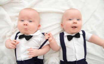 Two adorable twin baby boys dressed in matching white shirts, navy blue suspenders, and black bow ties lying on a soft white blanket. One baby is playfully pulling the other’s suspender