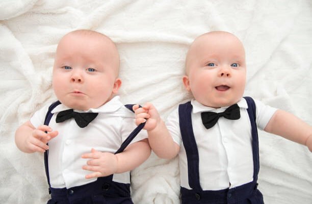 Two adorable twin baby boys dressed in matching white shirts, navy blue suspenders, and black bow ties lying on a soft white blanket. One baby is playfully pulling the other’s suspender